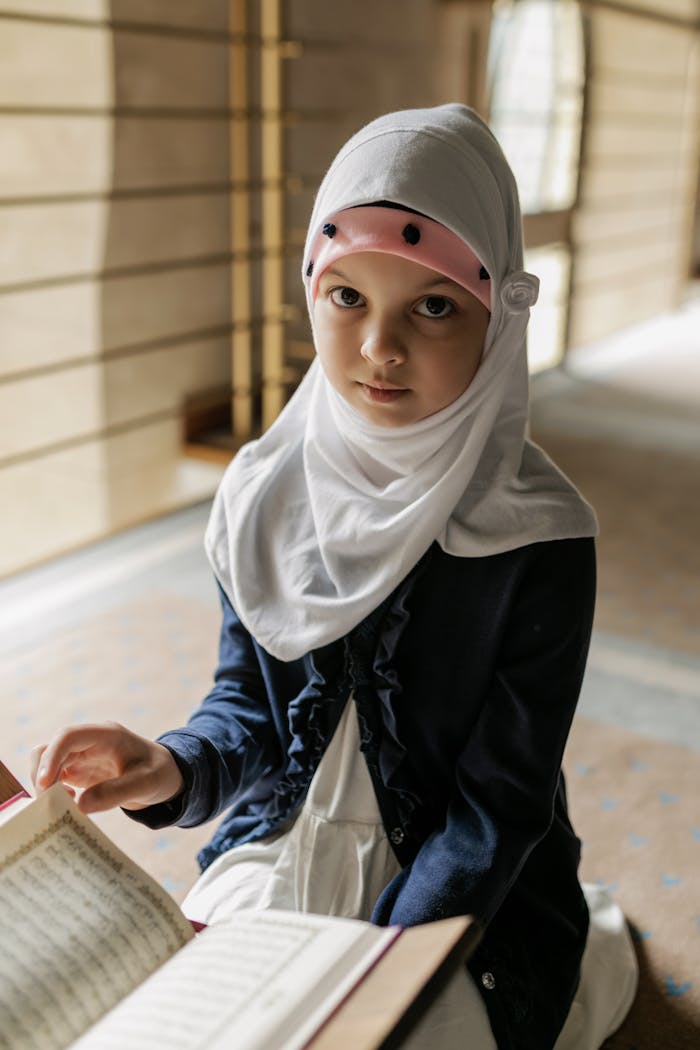 Portrait of a young girl in a hijab reading the Quran indoors. The scene captures a peaceful, cultural moment.